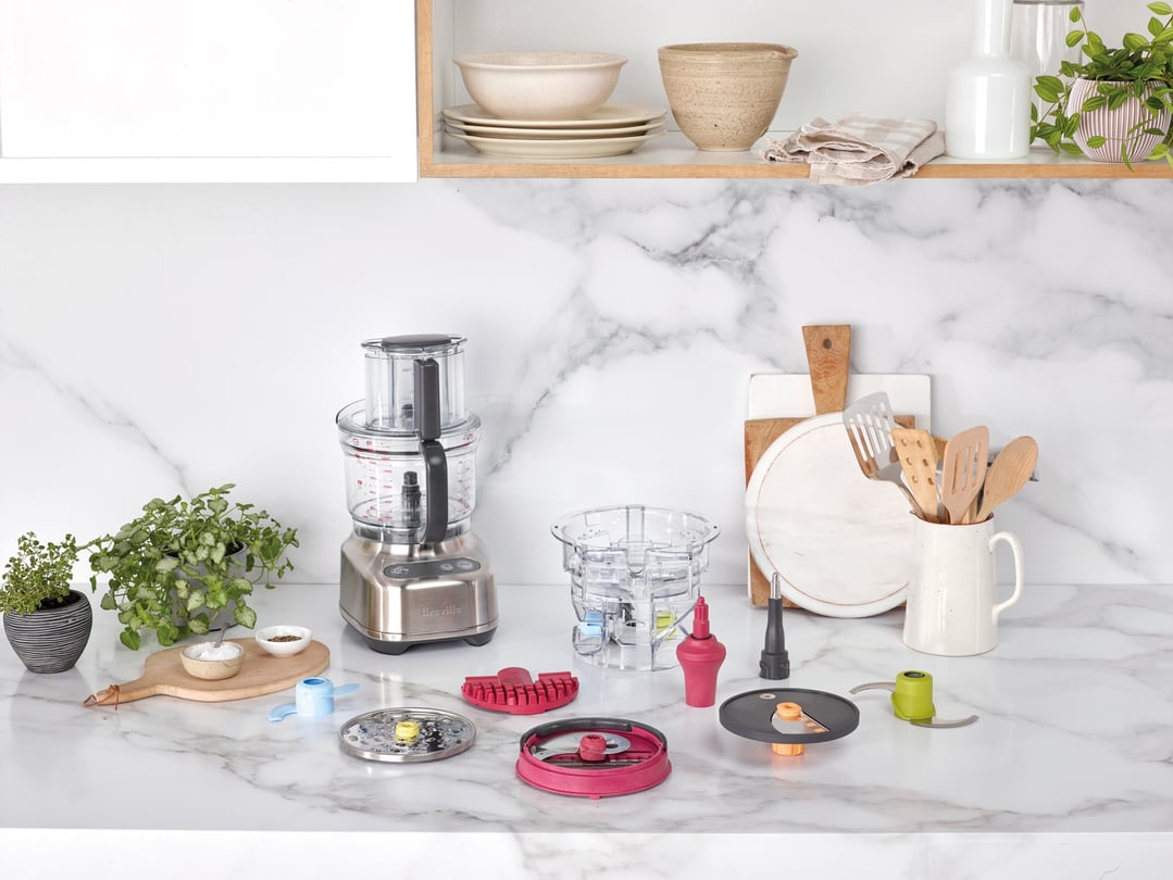 Breville food processor with accessories on a marble kitchen counter with plants