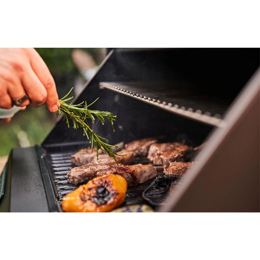 Hand adding rosemary to meat and vegetables cooking on a portable electric BBQ