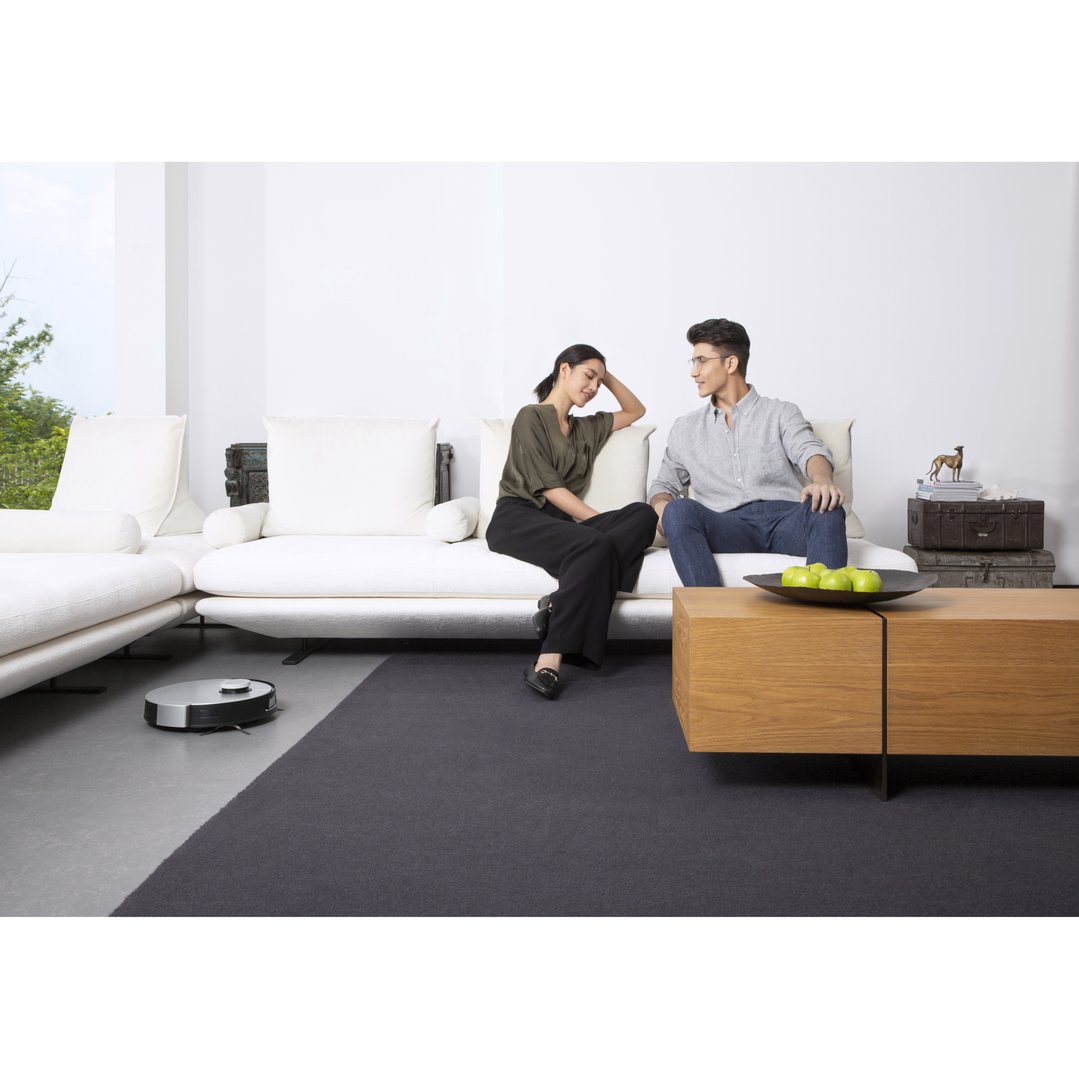 A robotic vacuum cleaning the floor in a living room with two people on a sofa