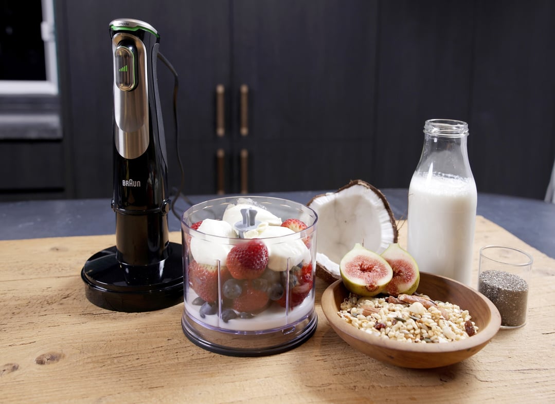 Hand blender on a wooden surface with fruit, milk, and cereal