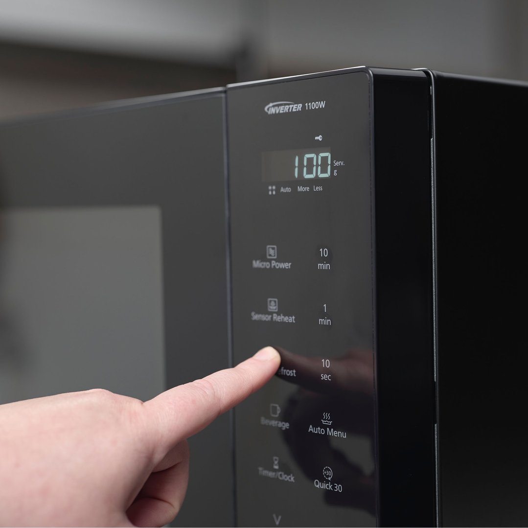 A hand presses the defrost button on a black Panasonic microwave
