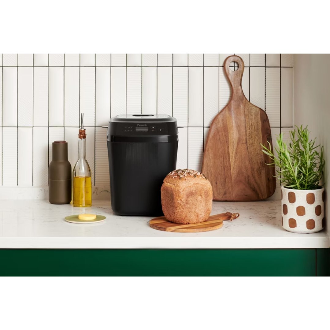 Black Panasonic bread maker on a kitchen counter with fresh bread and oil