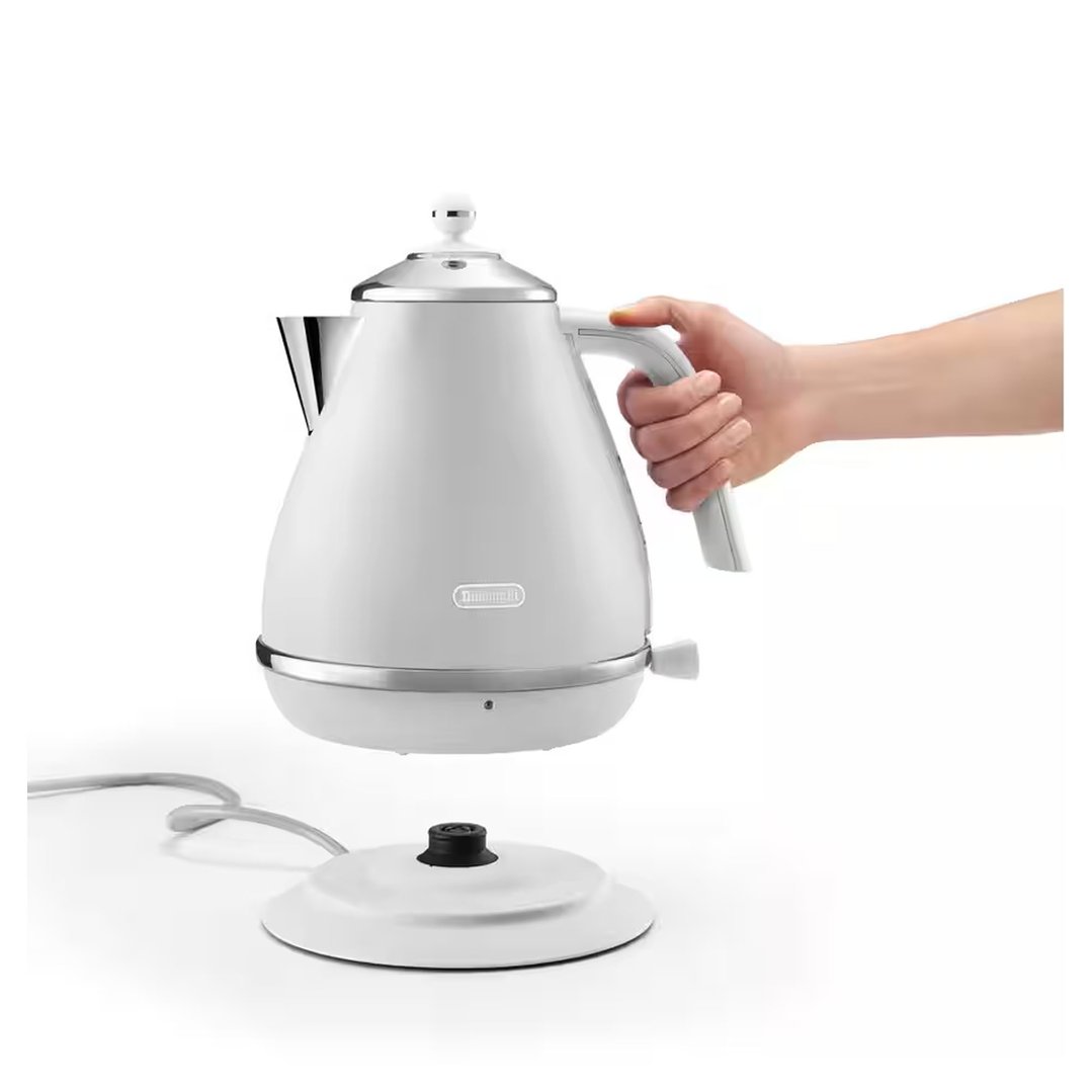 Hand holding a white kettle above its base on a white surface
