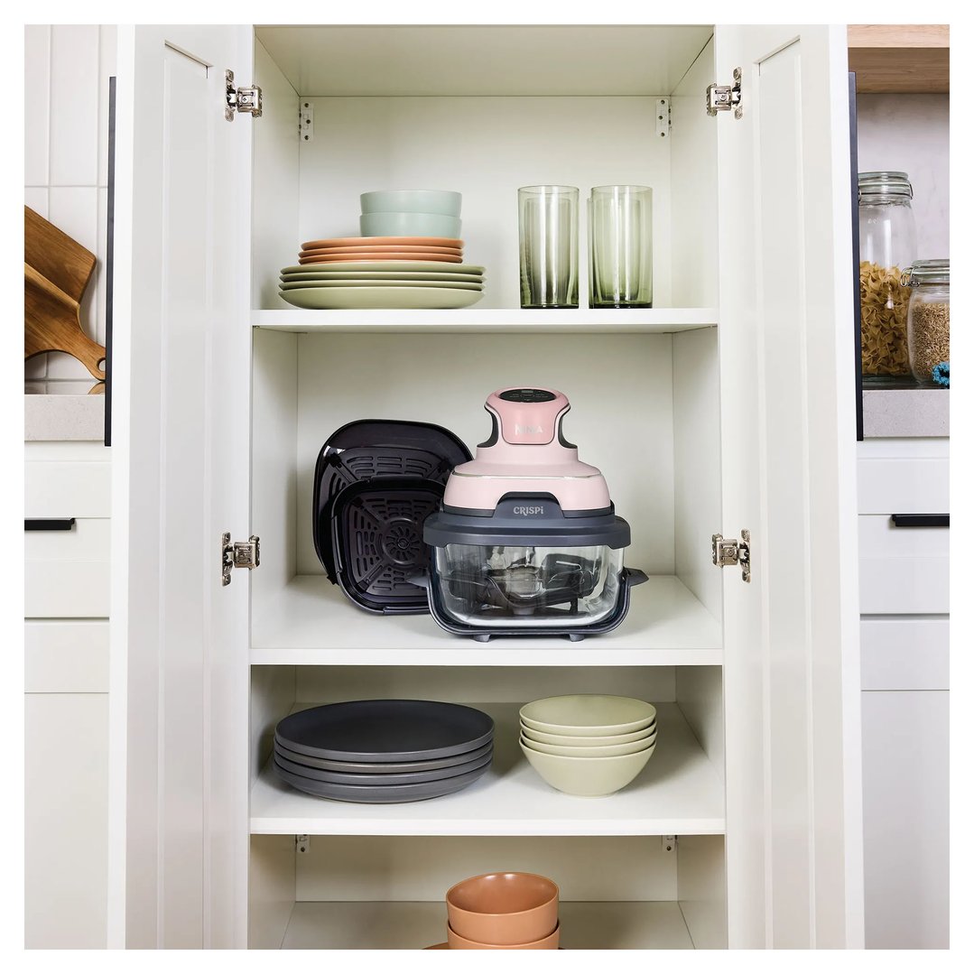 Pink Crispi air fryer and accessories on a shelf in a white kitchen cabinet