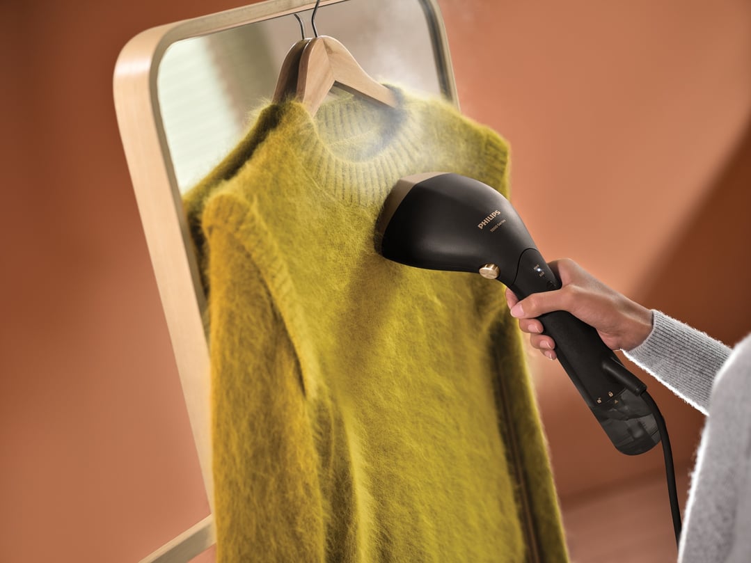 Handheld garment steamer steaming a yellow sweater hanging on a wooden hanger