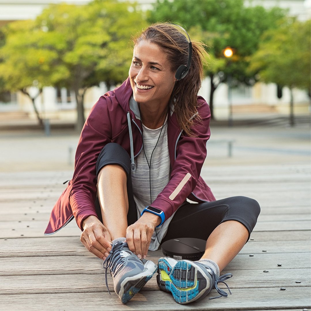 Woman sitting on a wooden deck, tying her shoe, with a portable CD MP3 player nearby