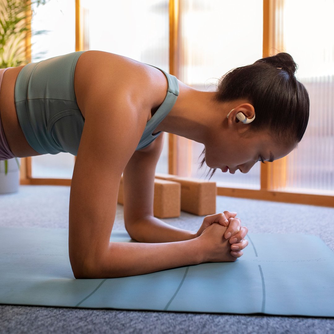 Woman in a plank position on a yoga mat wearing beige SHOKZ OpenFit headphones