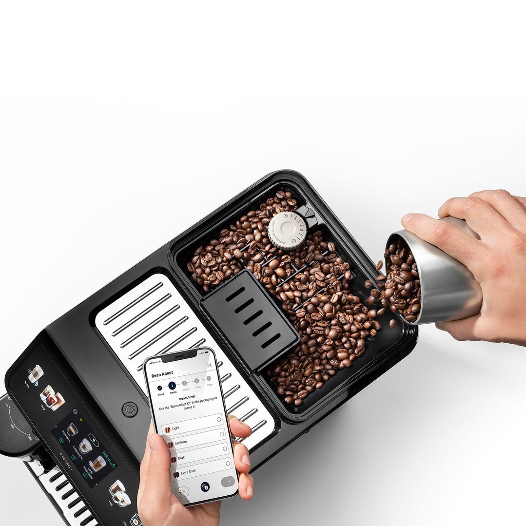 Hand pouring coffee beans into the Eletta Explore coffee machine, with phone app visible