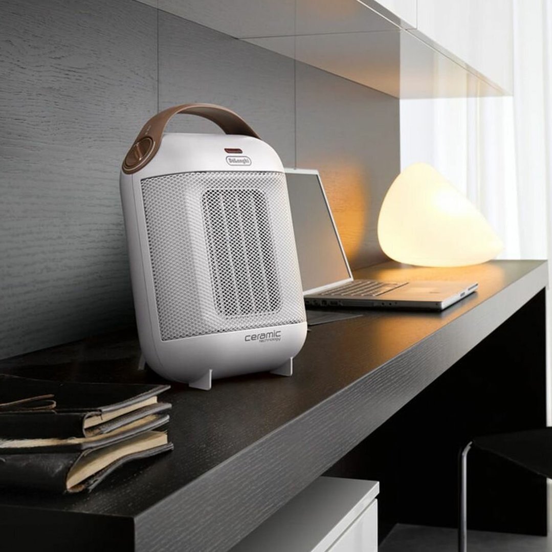 White ceramic heater on a dark desk with a laptop and lamp