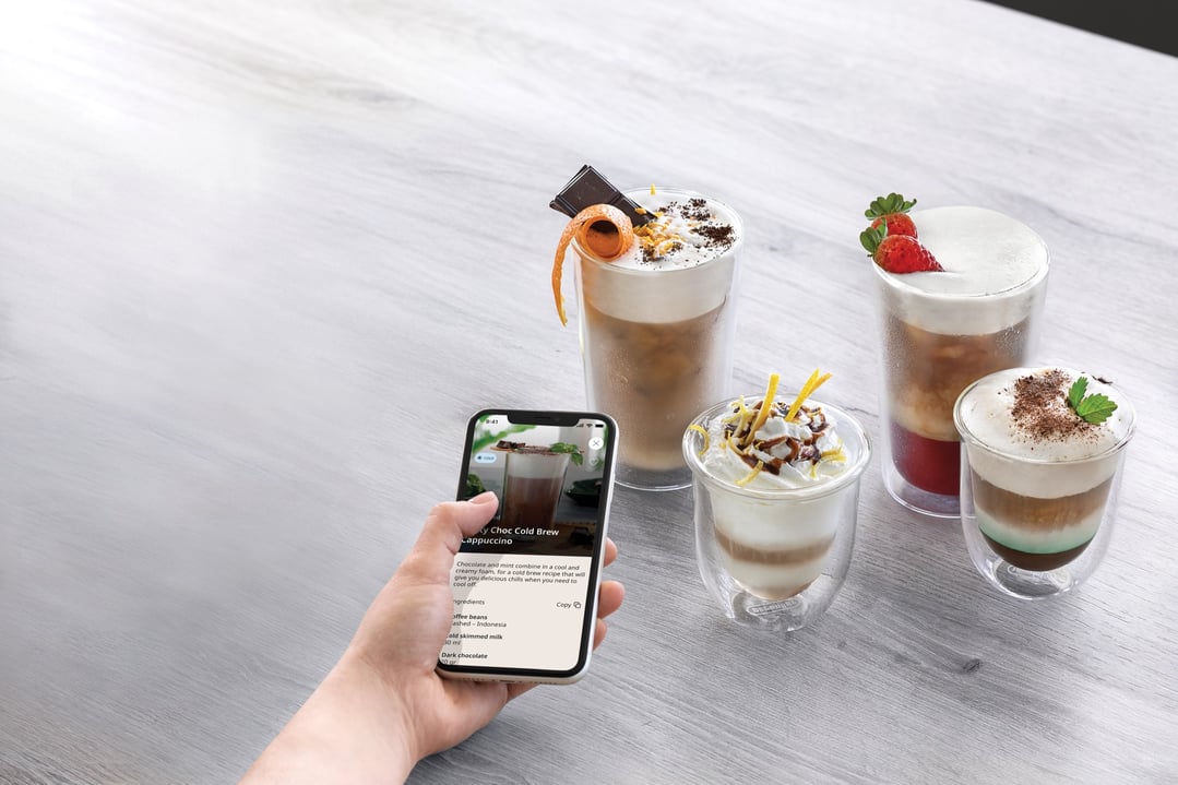 Hand holding a smartphone displaying a coffee recipe next to four coffee drinks