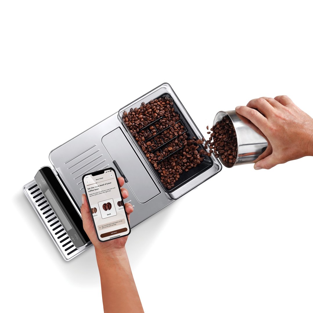 Hands pouring coffee beans into a coffee machine while holding a smartphone