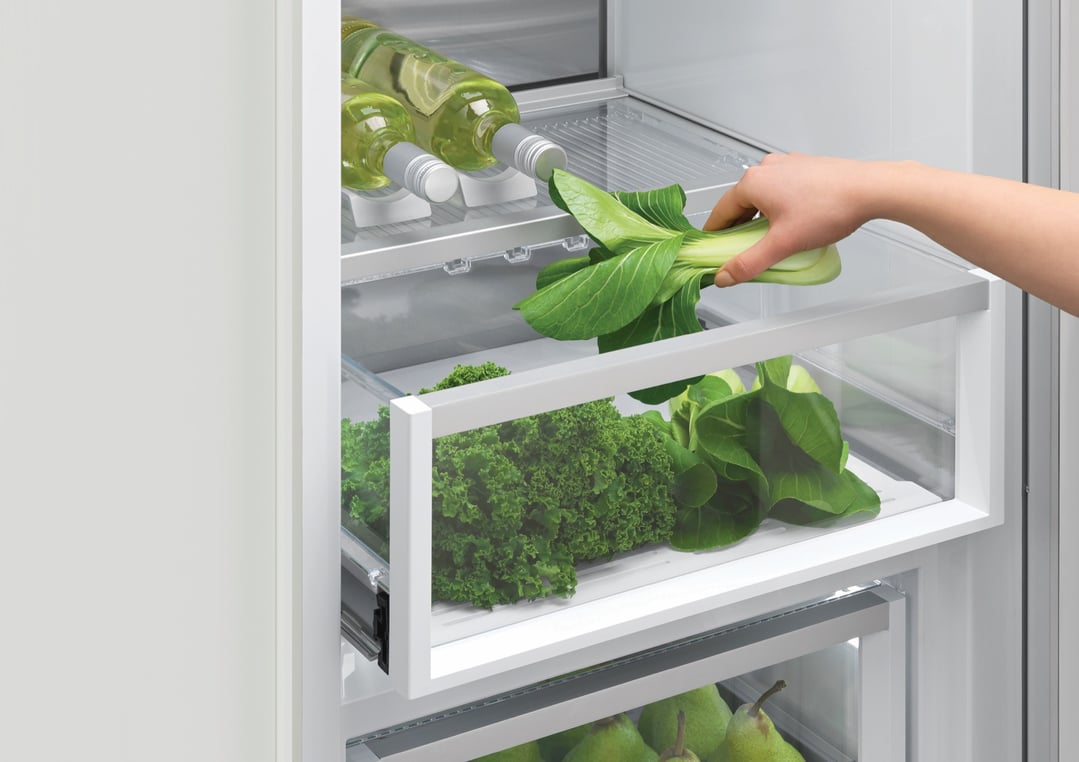 Hand placing bok choy into a refrigerator drawer with other vegetables and wine