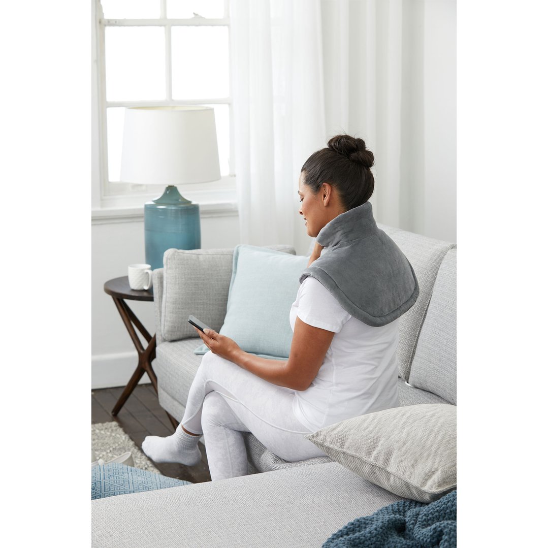 Woman wearing a gray heating pad on her shoulders, sitting on a sofa
