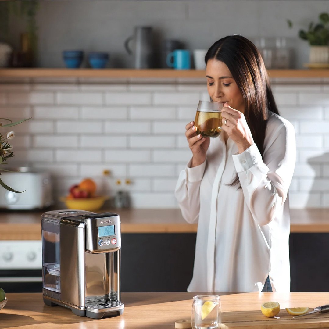 Woman drinking tea from a glass next to the Breville AquaStation Hot Water Dispenser