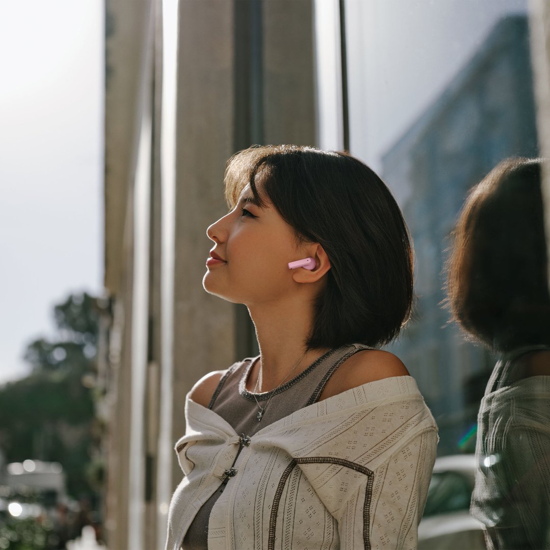 Woman wearing pink in-ear headphones, looking up in a sunlit outdoor setting