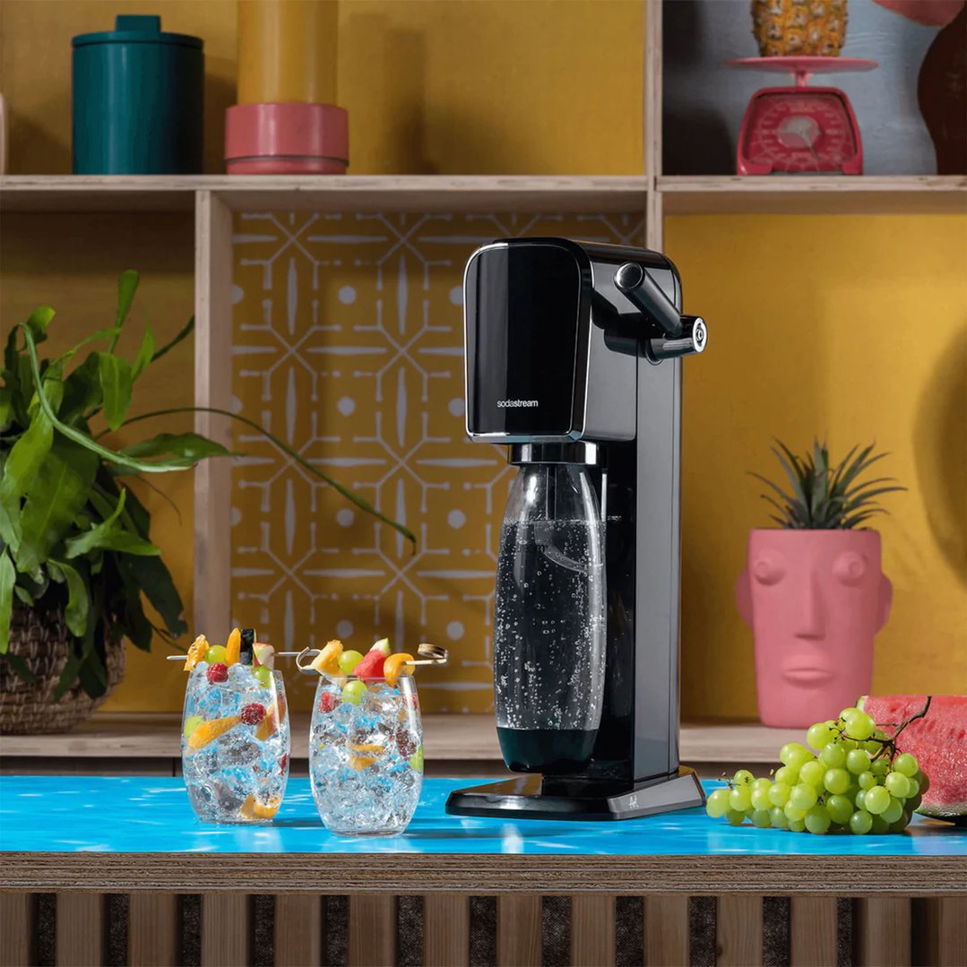 Black SodaStream Art Sparkling Water Maker on a blue counter with drinks and fruit