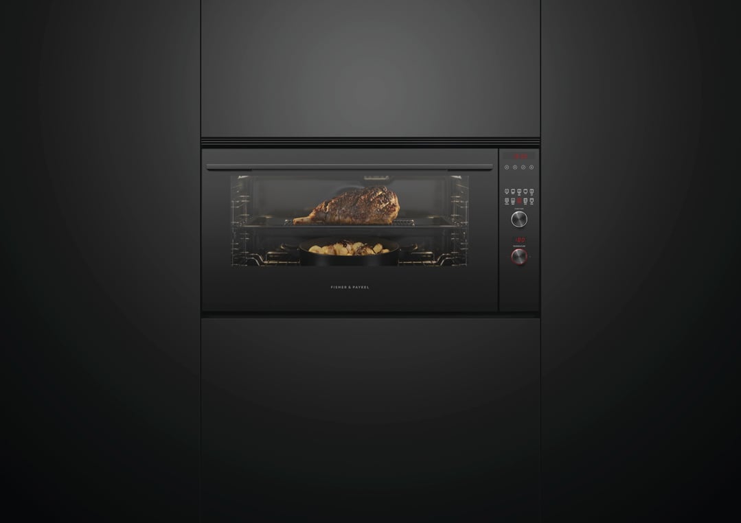 Black built-in oven cooking a leg of lamb and potatoes in a dark kitchen