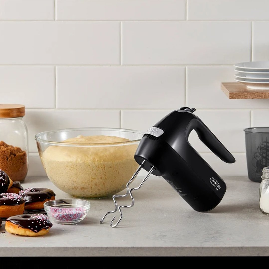 Black hand mixer with dough hooks next to a bowl of dough and donuts