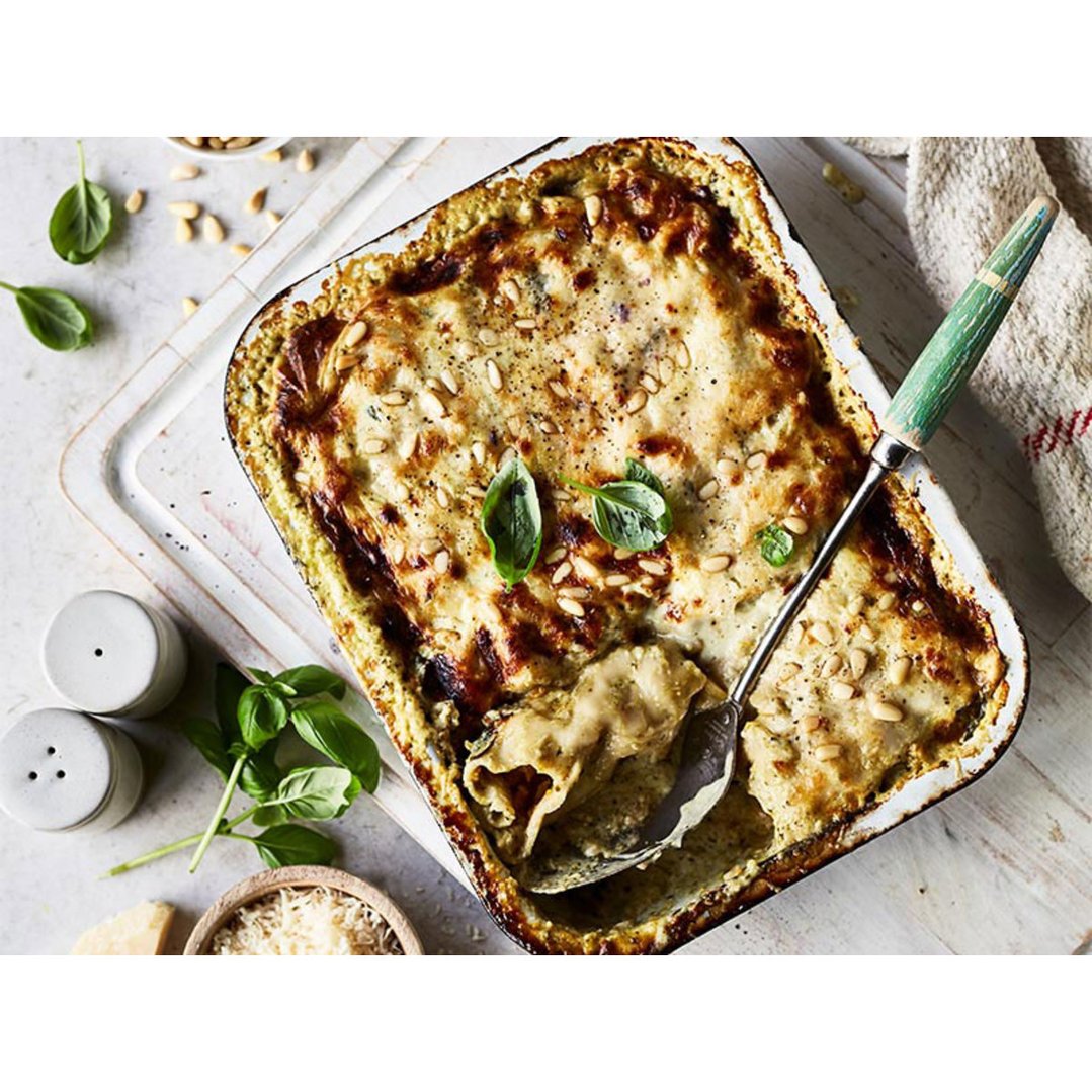 Lasagne in a baking dish with a serving spoon, basil, and pine nuts