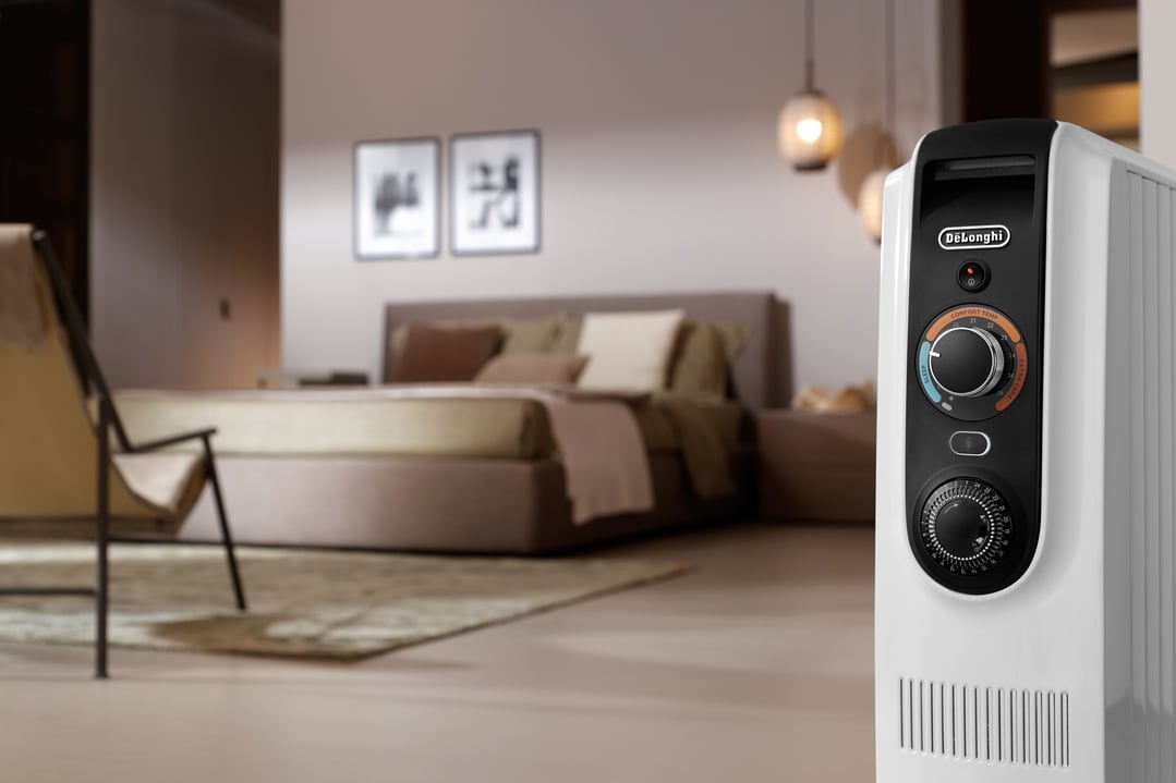 DeLonghi oil column heater in a bedroom with a bed and chair in the background