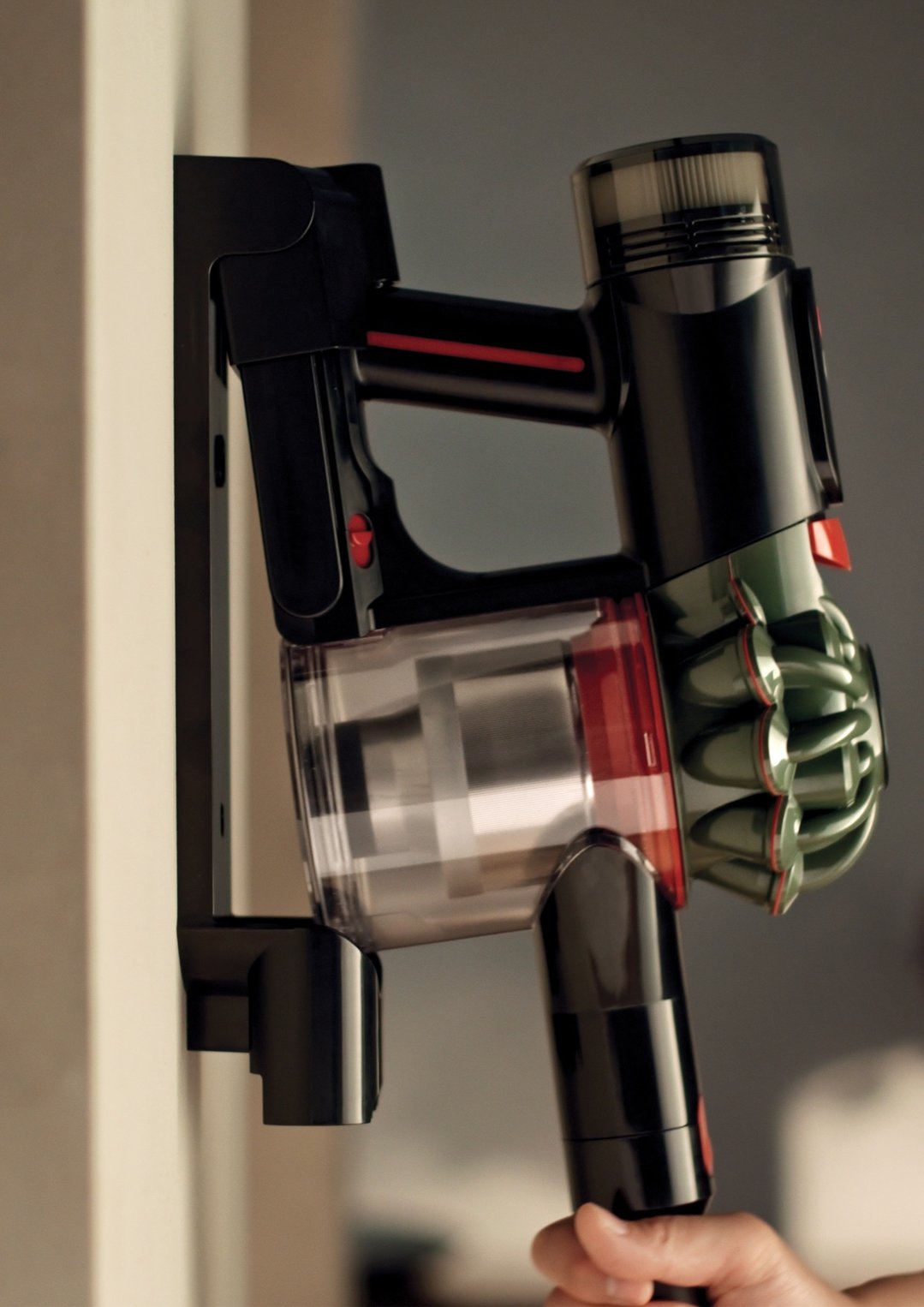 Hand holding a black and red handstick vacuum cleaner mounted on a wall