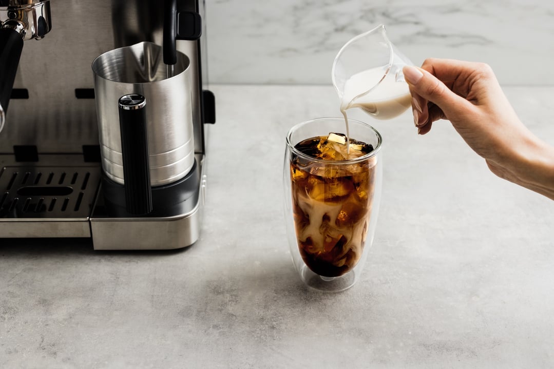 Hand pouring milk into a glass of iced coffee next to a coffee machine