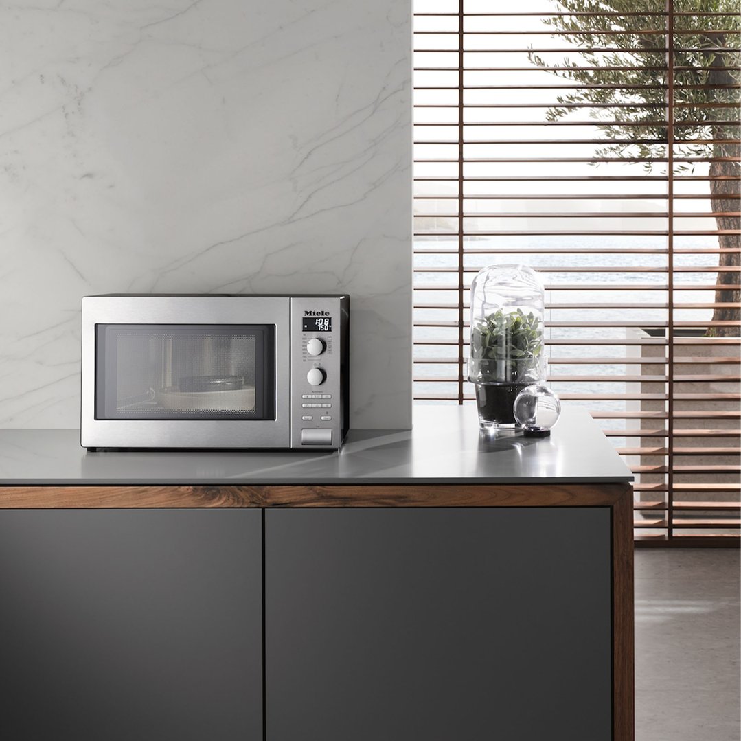 Stainless steel microwave oven on a kitchen counter with a plant and window blinds
