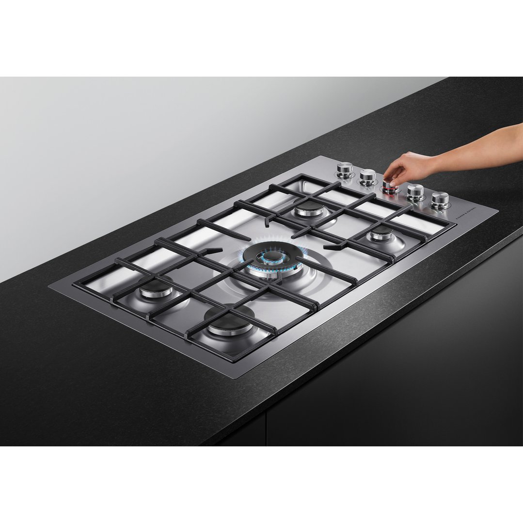 Hand adjusting control knob on a five burner gas cooktop with one burner lit