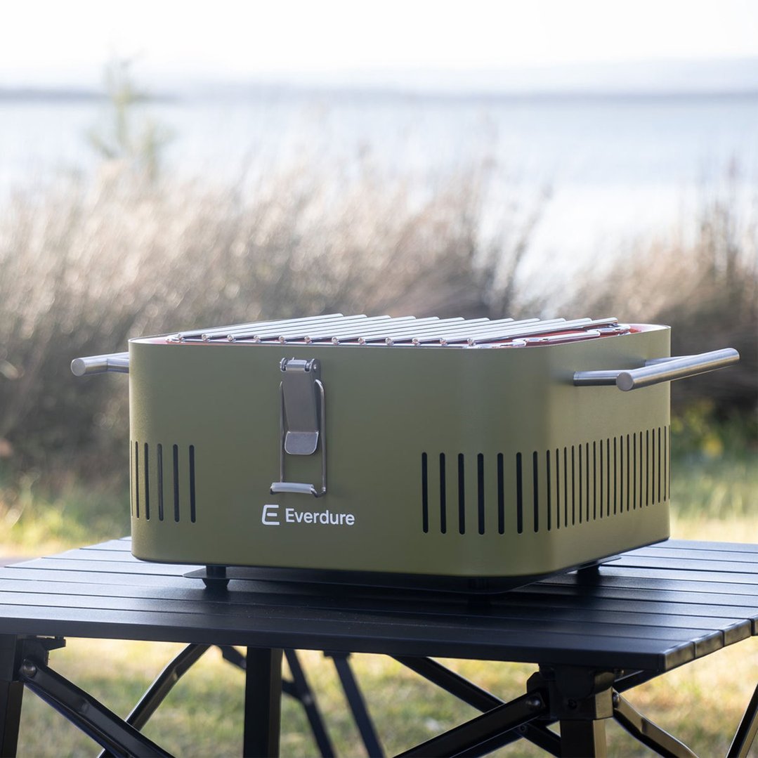 Khaki portable charcoal barbeque on a black table outdoors with a lake background