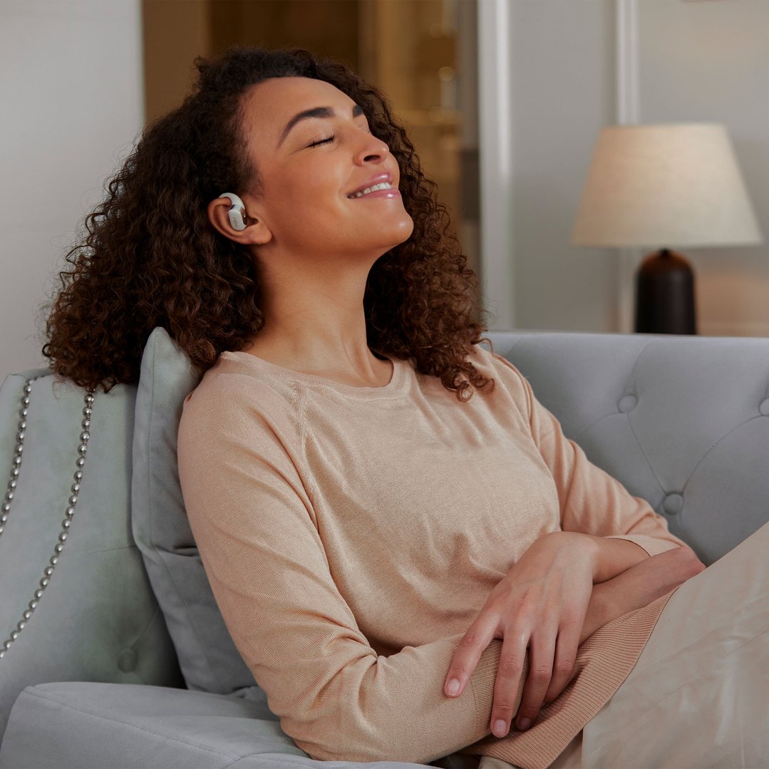 Woman wearing beige OpenFit headsets, relaxing on a grey sofa with eyes closed