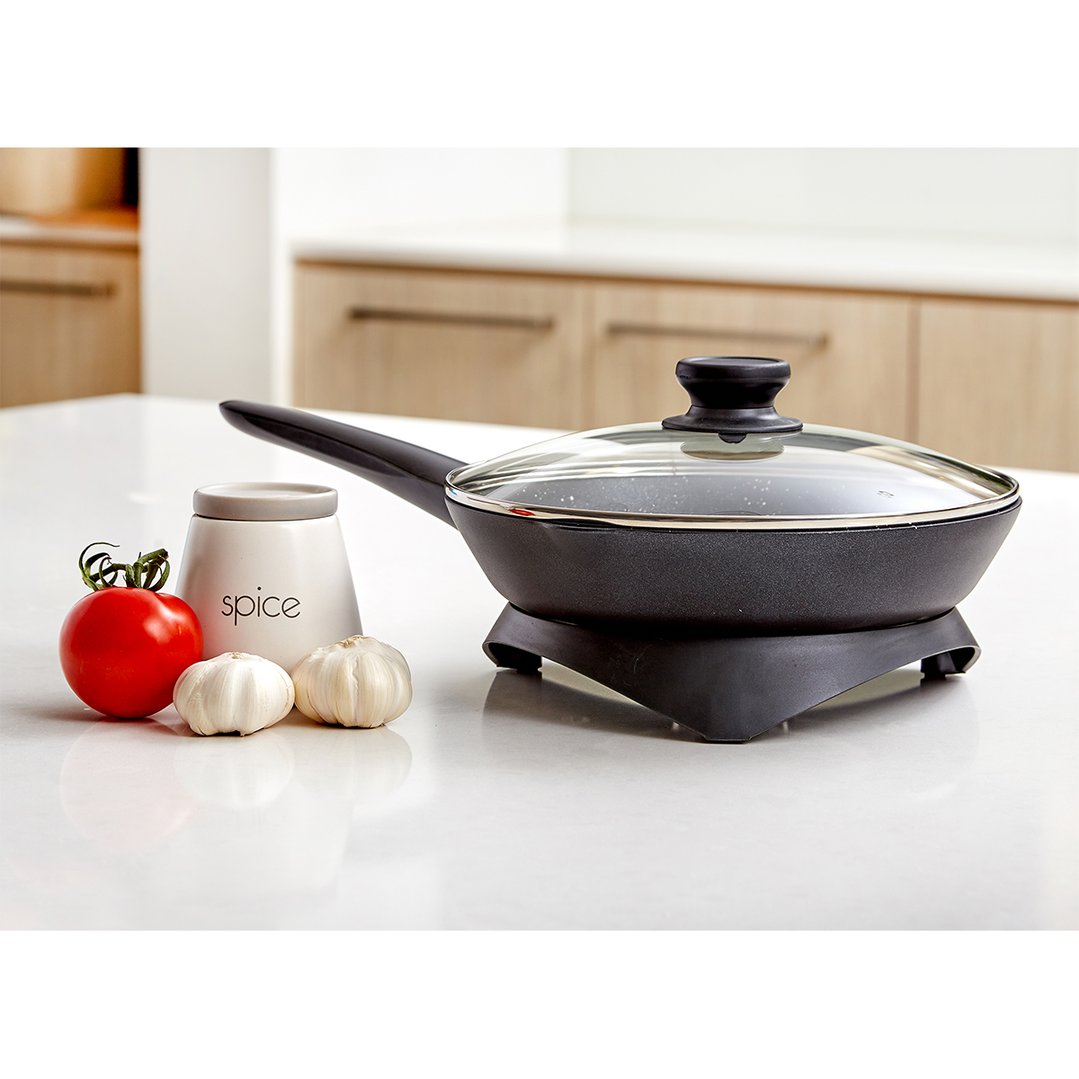 Electric skillet with lid on a white counter with tomato, garlic, and spice jar