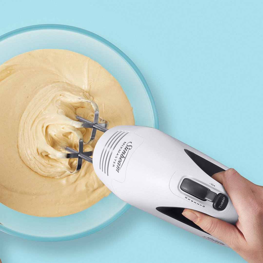 Hand holding a Sunbeam Mixmaster hand mixer mixing batter in a glass bowl