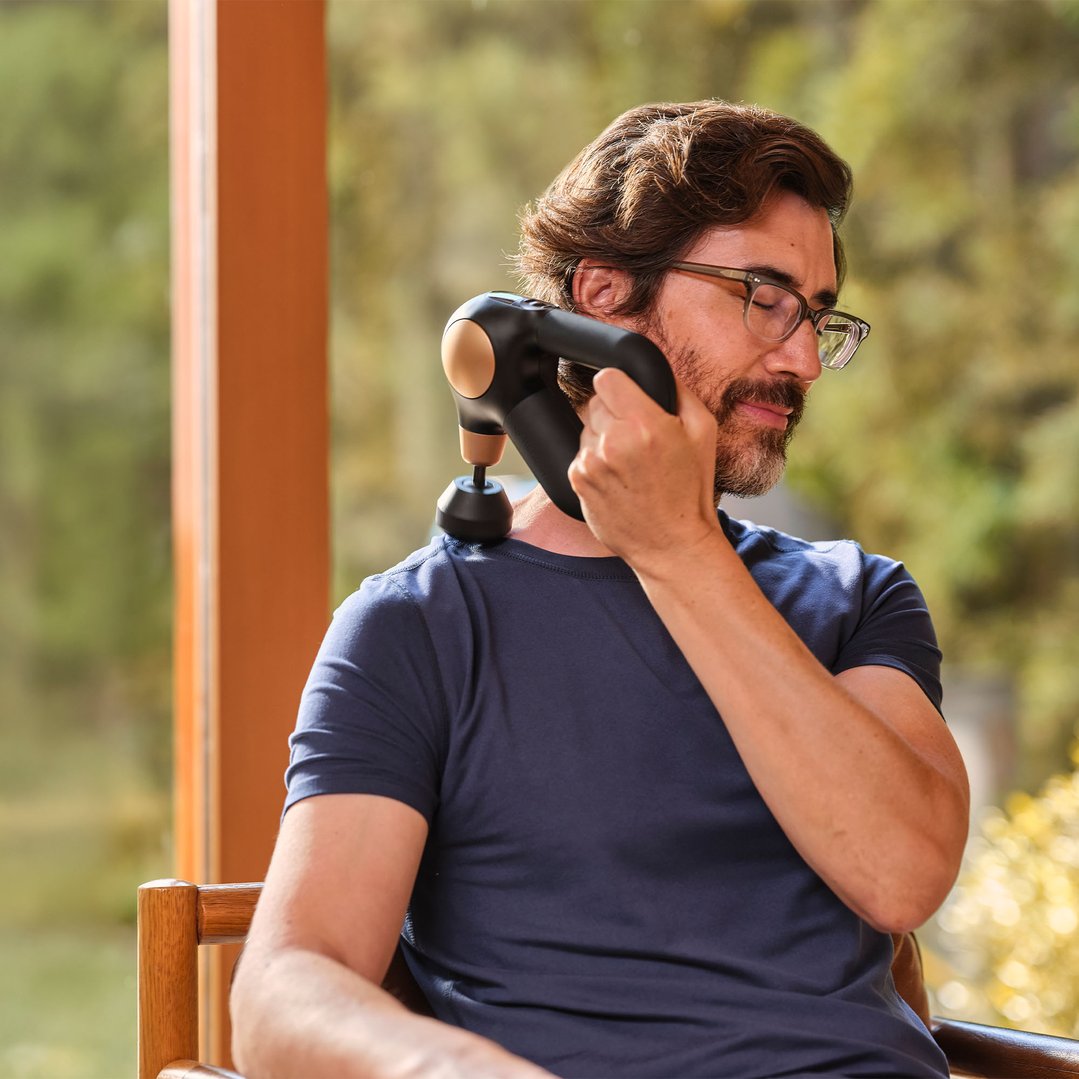 Man using a Theragun Sense massager on his shoulder while sitting outdoors
