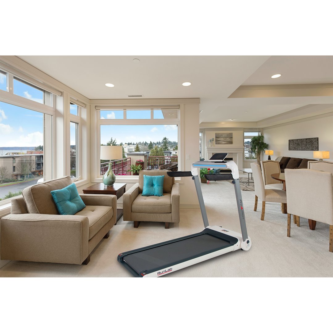 A treadmill in a modern living room with large windows and comfortable seating