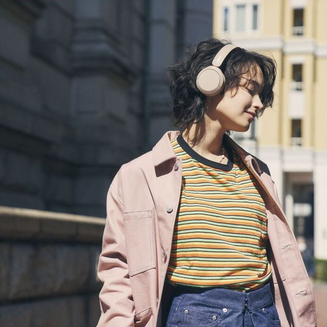 Woman wearing beige Sony wireless headphones outdoors, looking down and smiling