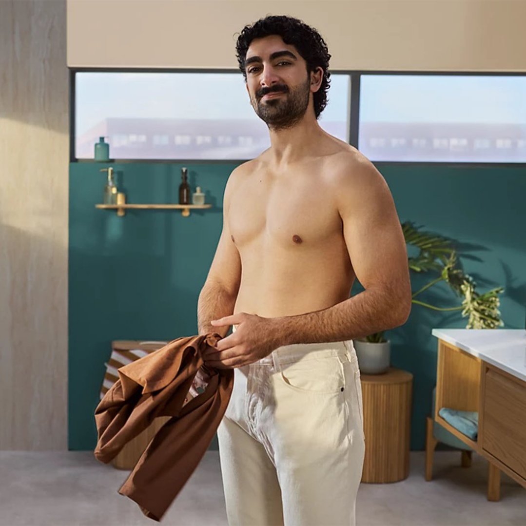 Man with a beard and curly hair standing shirtless in a bathroom