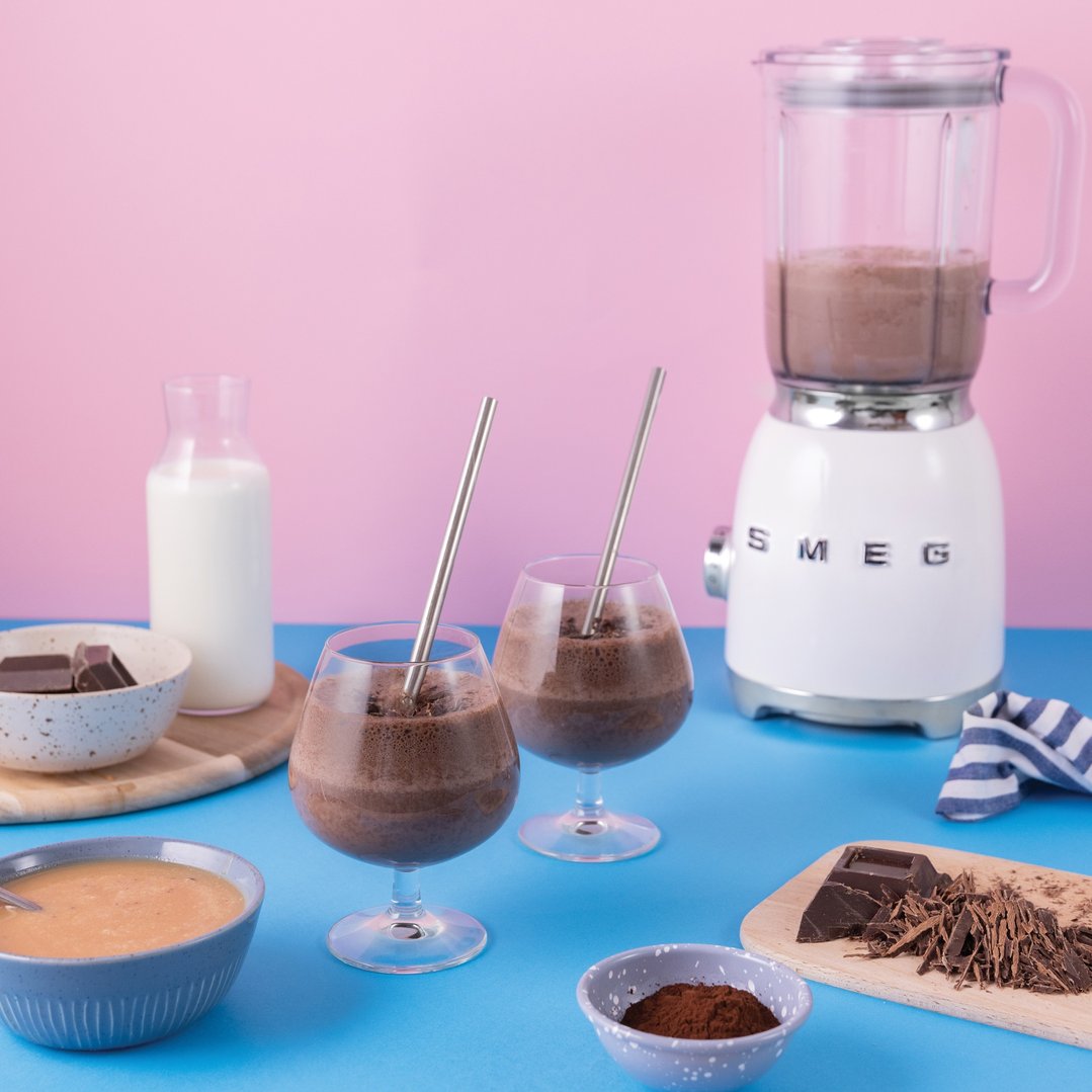 Two chocolate smoothies with ingredients and a Smeg blender on a blue surface
