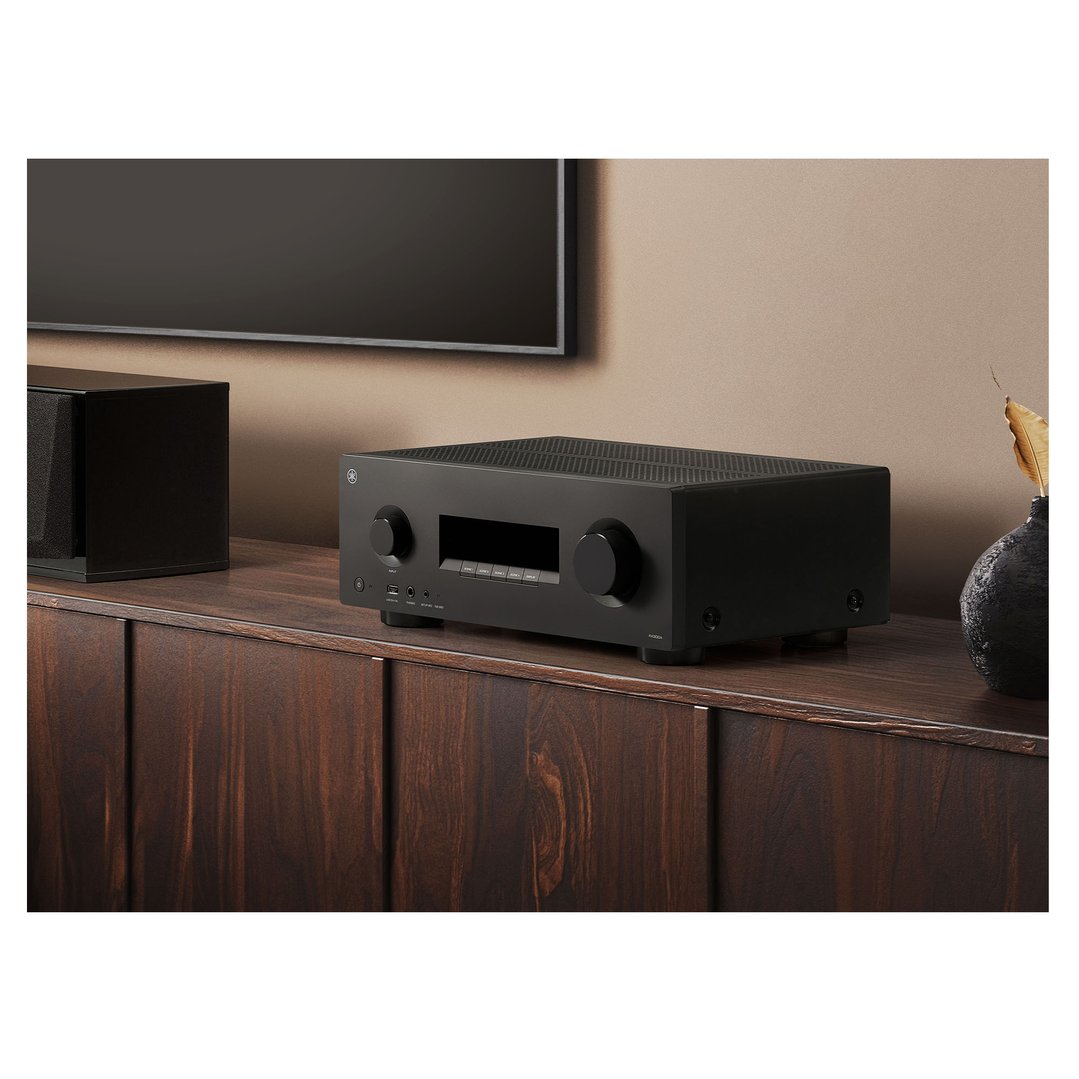 Black AV receiver on a wooden cabinet with a TV and speaker in the background