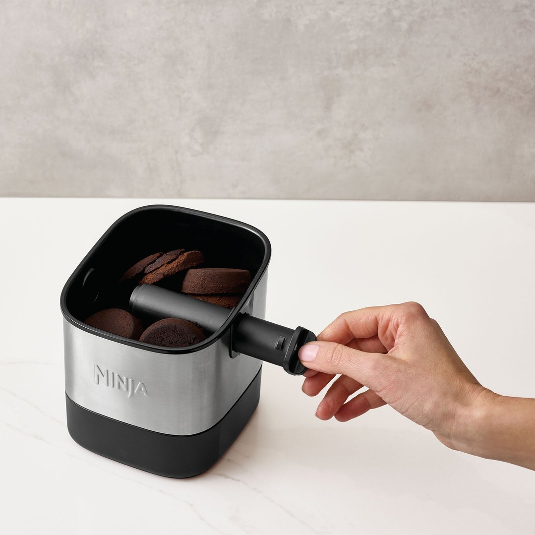 Hand operating the Ninja Luxe Café Knock Box filled with coffee grounds