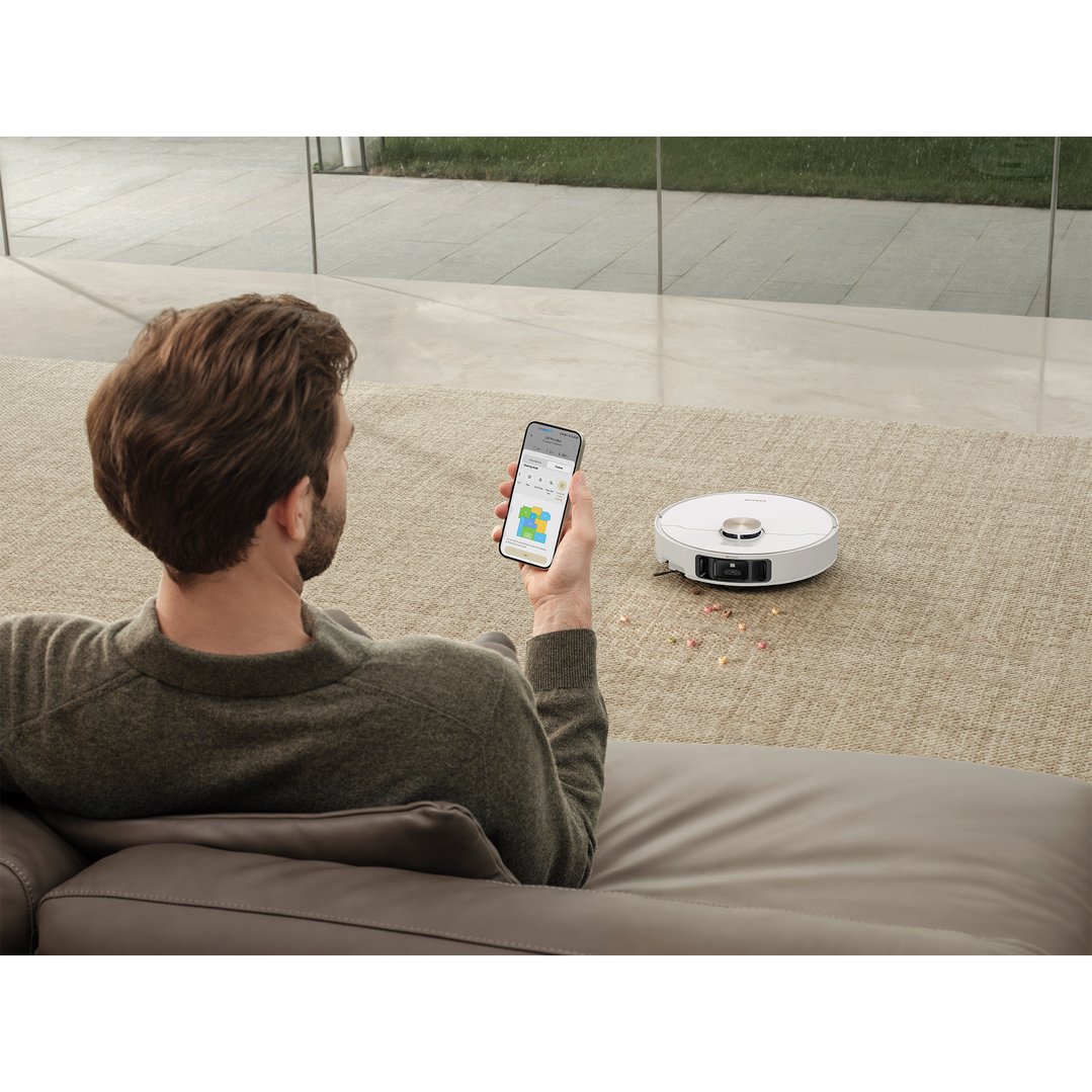 Man on sofa using phone to control a white robot vacuum cleaner on carpet