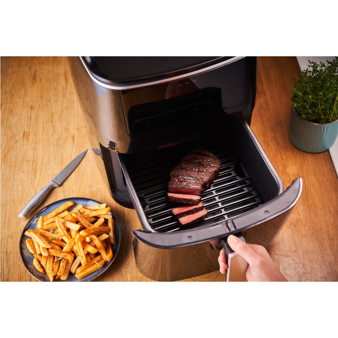 Hand pulling out air fryer drawer with steak and fries on a wooden counter