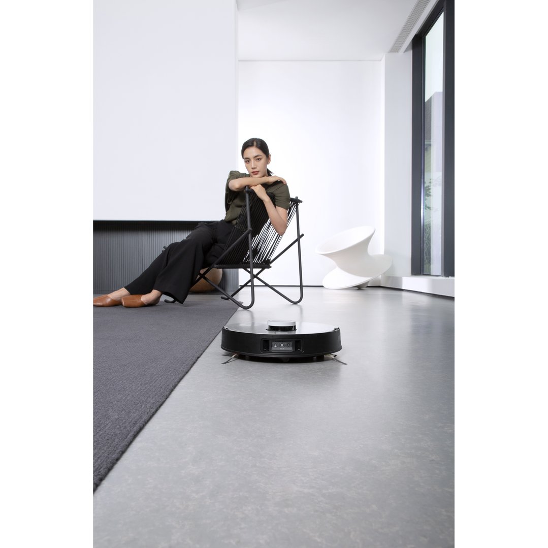 A robotic vacuum on a grey floor with a woman sitting in a chair nearby