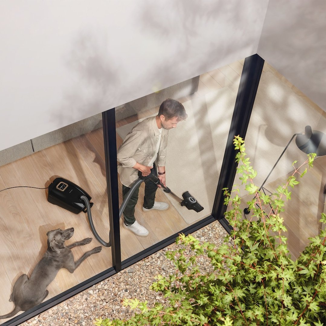 Man vacuuming a rug with a Miele vacuum cleaner, dog lying nearby