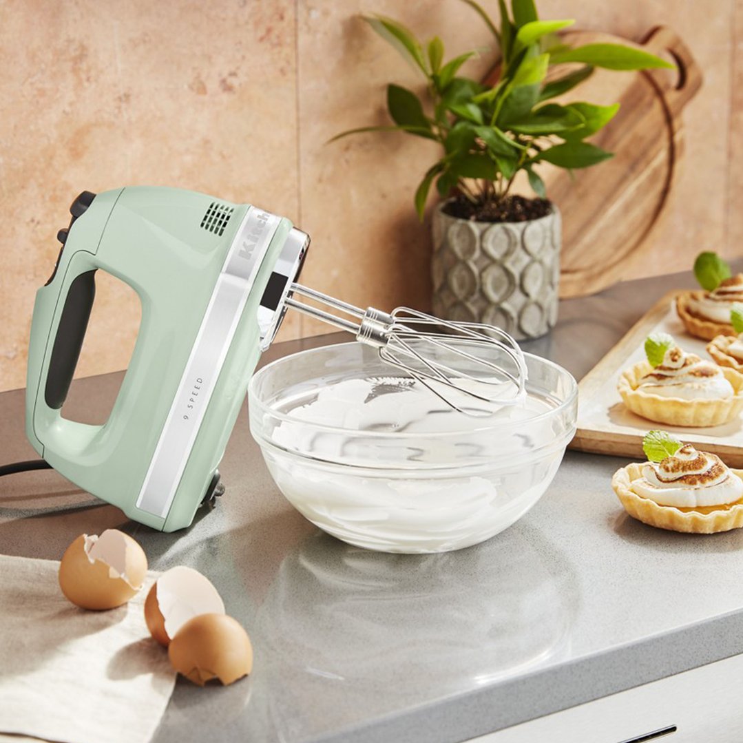 Pistachio KitchenAid hand mixer with whisk attachments mixing meringue in a bowl on a counter