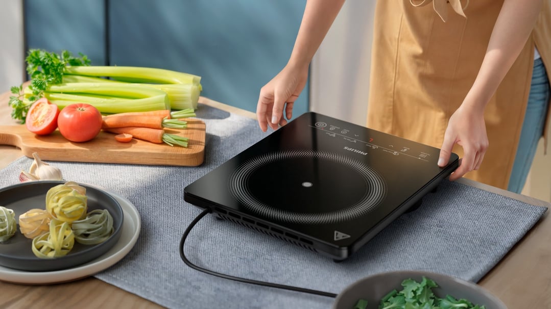 Person's hands adjusting a black induction cooker on a table with vegetables