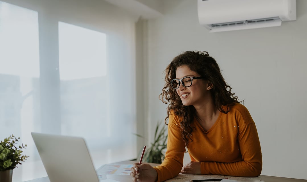 Woman with glasses smiling and writing at a desk with a laptop