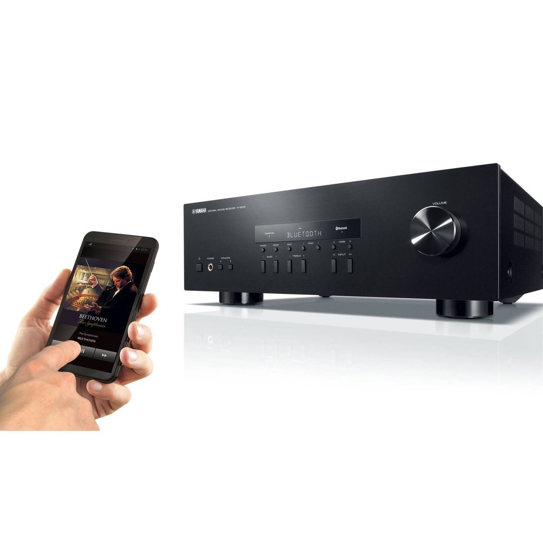 Hand holding a smartphone displaying music, next to a black Yamaha HiFi receiver