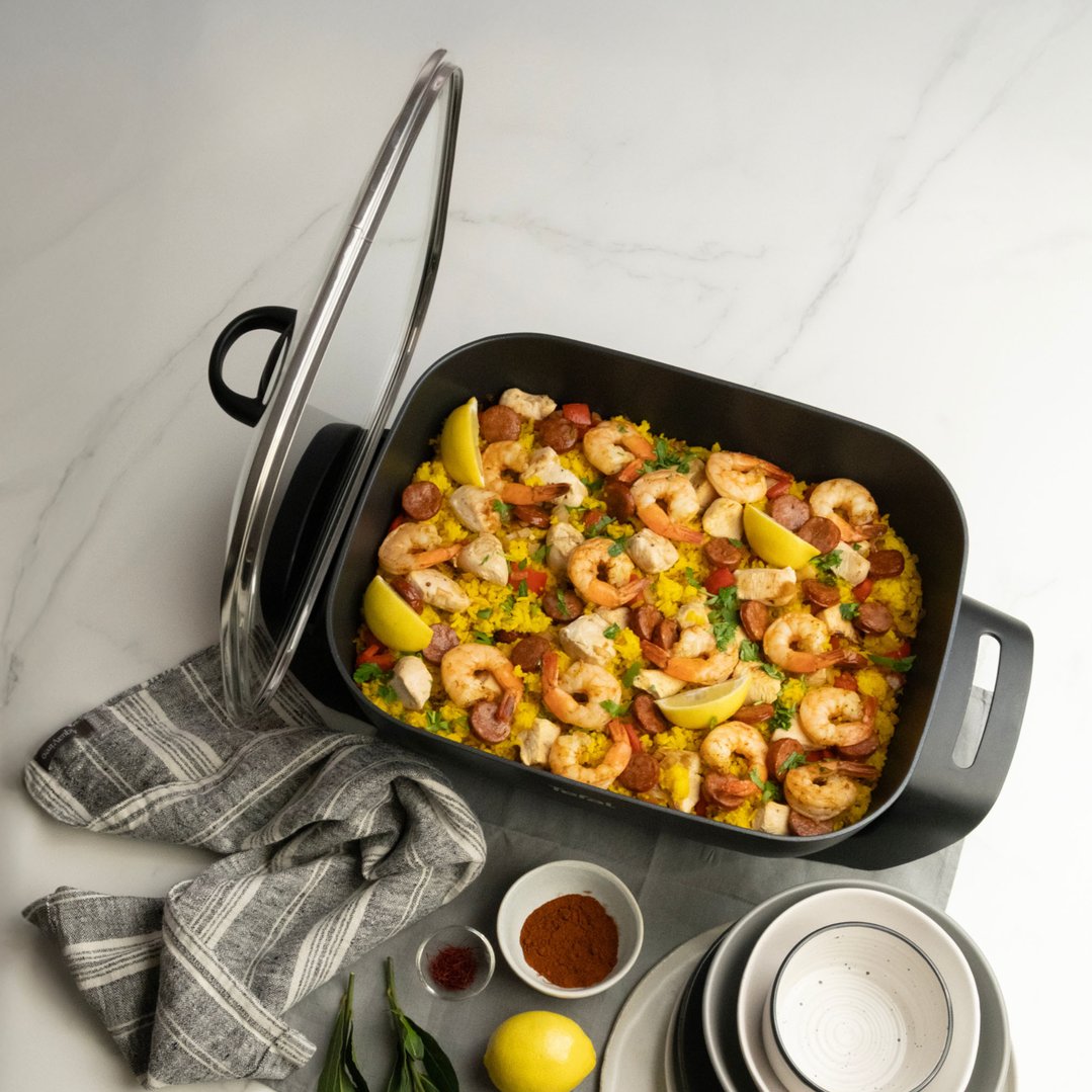 Electric frypan filled with paella, surrounded by ingredients and dinnerware on a counter