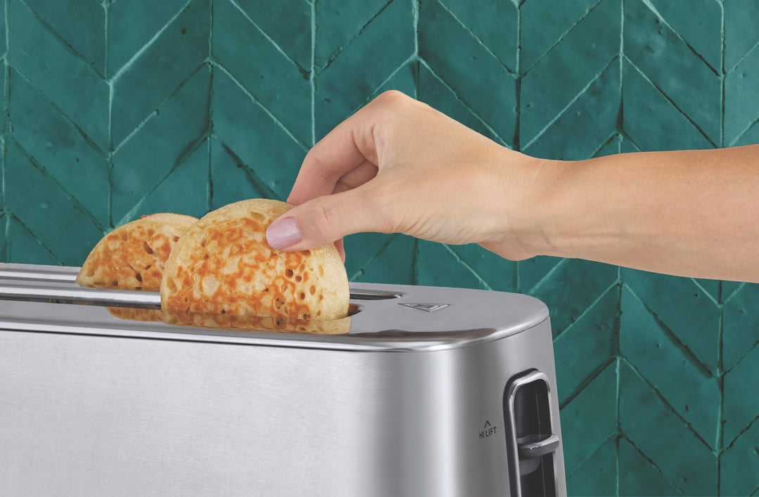 Hand placing crumpets into a stainless steel toaster against a teal tiled wall
