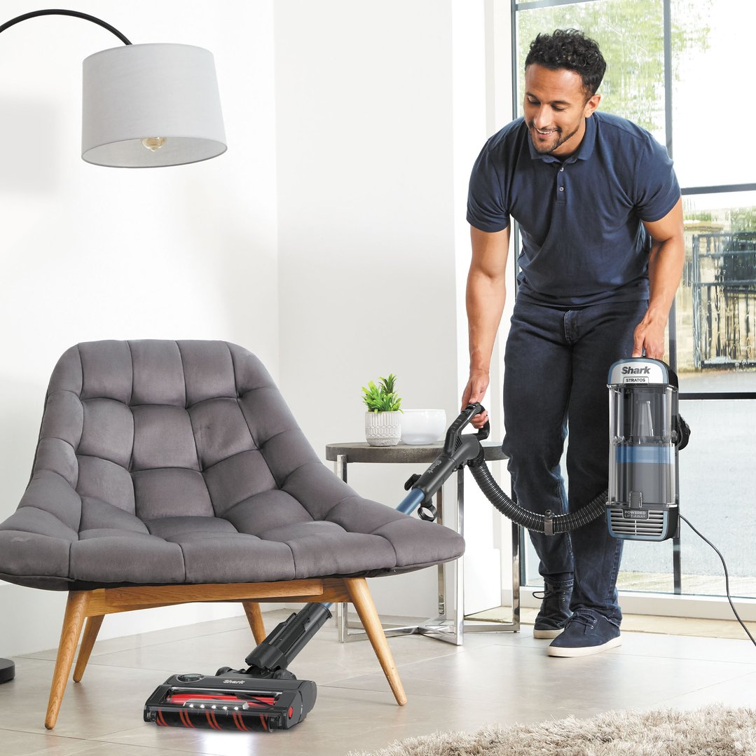 Man vacuuming around a gray armchair with a Shark Stratos vacuum cleaner